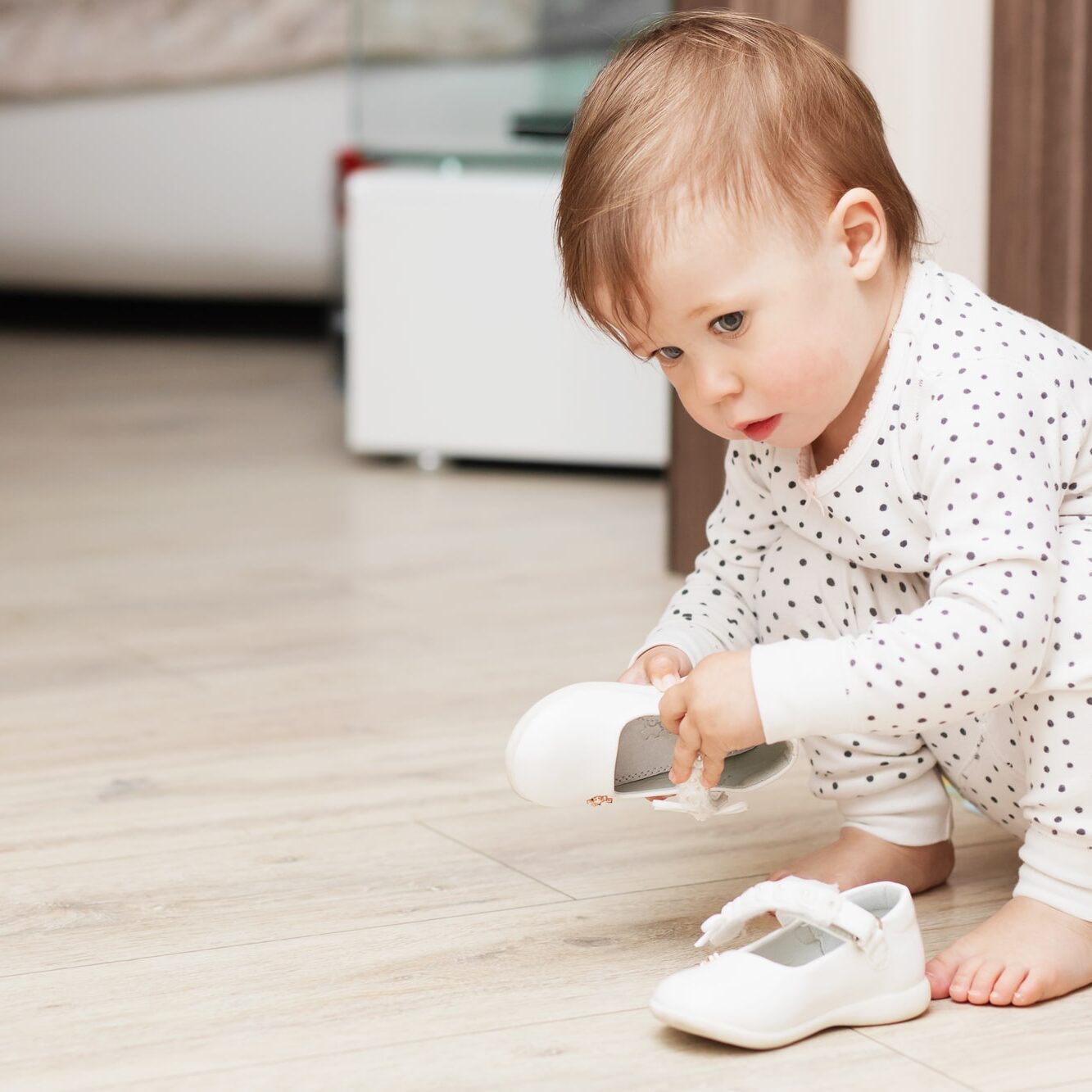 Cute baby girl in pajamas squatting on floor tries to put on her white shoes in the playroom